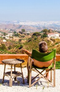 Mann sitzt in der Sonne in einem grünen Stuhl auf einer Bergterrasse und sieht entspannt über die hügelige Landschaft Spaniens. | © pexels.com|anna-m