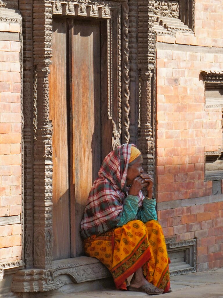 Eine ältere Frau sitzt in traditioneller nepalesischen Kleidung auf der Schwelle einer kunstvoll geschnitzten Eingangstüre direkt an der Straße. | © Constanze Schwarz