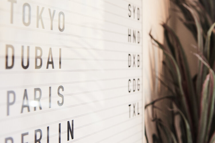 Eine Tafel mit Reisezielen hängt vor einer Grünpflanze an der Wand. Man sieht von der Seite darauf. | © Michael Zimmermann | BMZ Design Co.