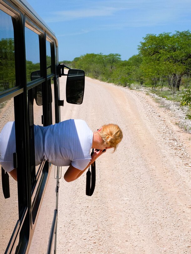 Eine Touristin lehnt sich während einer Safari mit der Fotokamera aus dem Fenster eines Busses um ein Bild zu machen. | © Manuela Holert